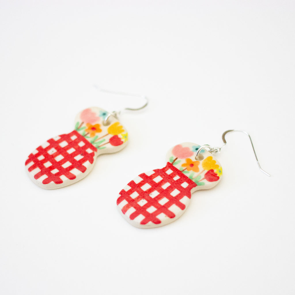 A pair of handmade ceramic earrings featuring a double-rounded shape, flowers in a vase. The top section is decorated with hand-painted colourful flowers, while the bottom section has a bold red checkered pattern. Finished with silver hooks.