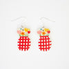 Front view of a pair of handmade ceramic earrings featuring a double-rounded shape, flowers in a vase. The top section is decorated with hand-painted colourful flowers, while the bottom section has a bold red checkered pattern. Finished with silver hooks.