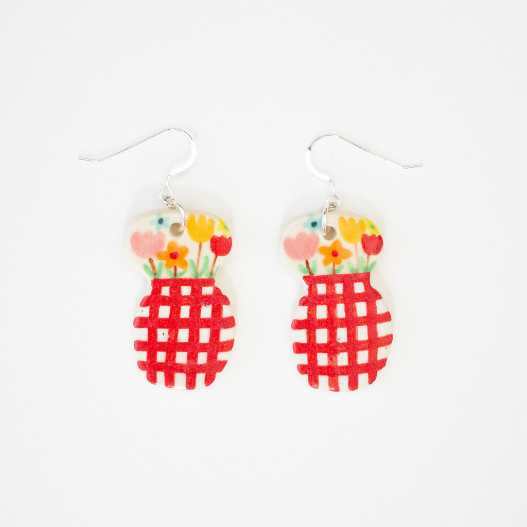 Front view of a pair of handmade ceramic earrings featuring a double-rounded shape, flowers in a vase. The top section is decorated with hand-painted colourful flowers, while the bottom section has a bold red checkered pattern. Finished with silver hooks.