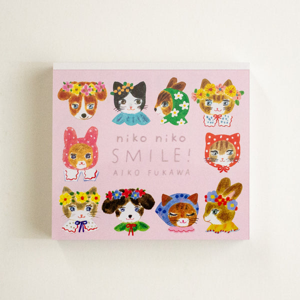 A square pink memo pad titled 'niko niko SMILE!' by Aiko Fukawa, featuring cute illustrated animals in colourful floral outfits on a light background.