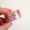 A hand holding a roll of pink washi tape featuring cute cartoon cat illustrations and small gold flower and bow accents, set against a plain off-white background.