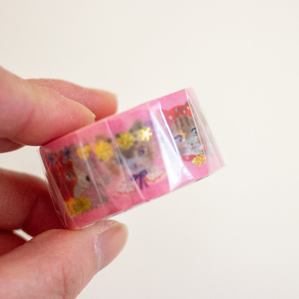 A hand holding a roll of pink washi tape featuring cute cartoon cat illustrations and small gold flower and bow accents, set against a plain off-white background.