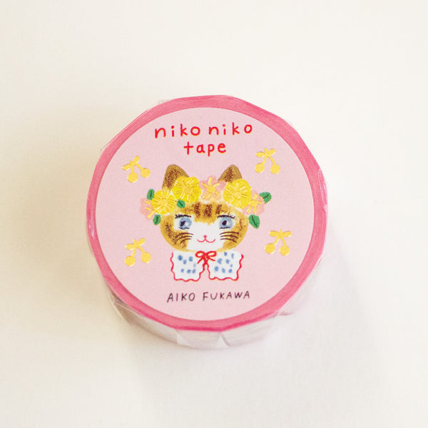 A roll of pink washi tape featuring a cute illustration of a cat wearing a yellow flower crown and a blue-and-white polka dot bow, with the text 'nikoniko tape' and 'AIKO FUKAWA' printed on the label.