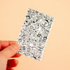 A hand holding a small white rectangular sticker with dense black doodle illustrations, against a plain warm beige background.
