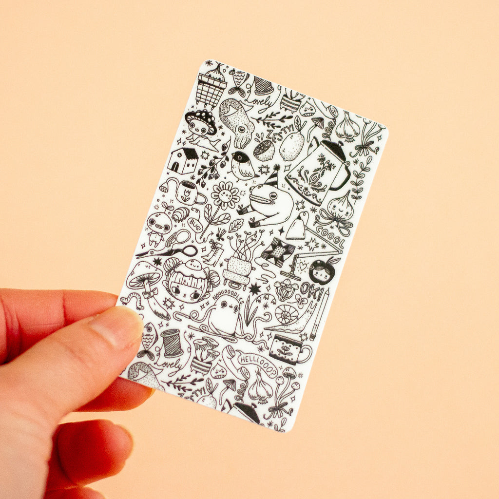 A hand holding a small white rectangular sticker with dense black doodle illustrations, against a plain warm beige background.
