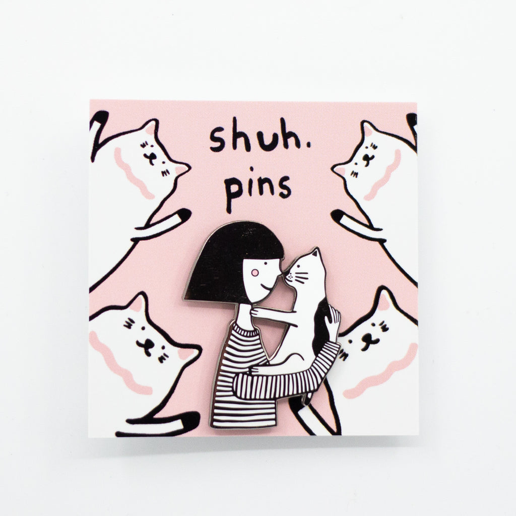 Enamel pin featuring two hand-drawn characters, a girl and her cat, touching noses in a kiss, displayed on a pink card with cat illustrations and the words “shuh. pins”.