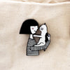 Enamel pin featuring two hand-drawn characters, a girl and her cat, touching noses in a kiss.