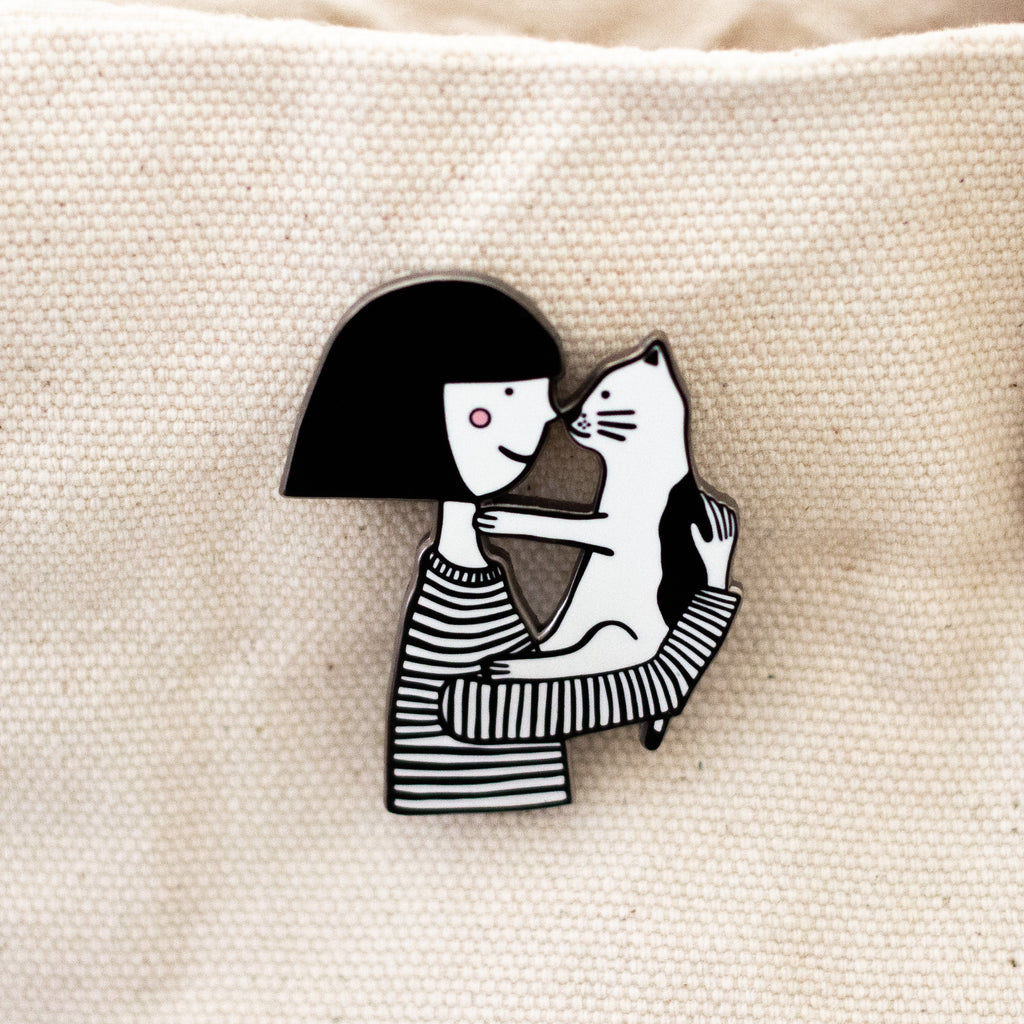 Enamel pin featuring two hand-drawn characters, a girl and her cat, touching noses in a kiss.