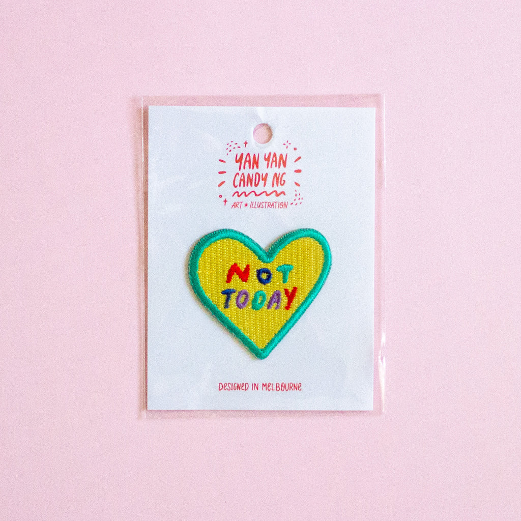 A heart-shaped embroidered patch with the text 'NOT TODAY' in colourful letters, outlined in green, displayed on a white backing card with a clear plastic sleeve against a solid pink background.