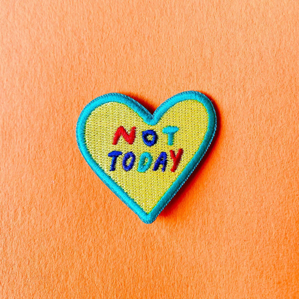 A heart-shaped embroidered patch with a yellow center, blue border, and the text 'NOT TODAY' in multicoloured letters, placed on a textured orange background.