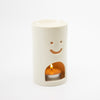 White ceramic oil burner with smiley face design featuring a lit candle inside on a white background