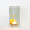 A green ceramic oil burner with a smiling face cutout and a tea light candle burning inside, set against a plain white background.