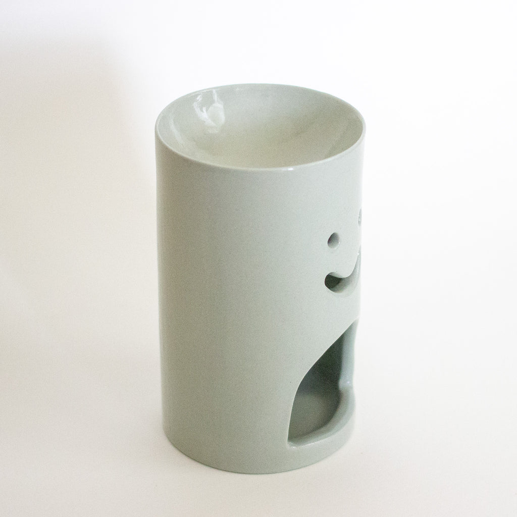 A sage green ceramic oil burner with a minimalist smiley face design and a bottom ventilation hole, set against a plain white background.