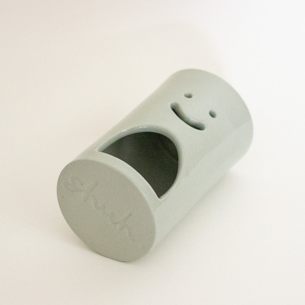 A green ceramic cylindrical container with a smiley face cutout on one end and a large arched opening on the side, photographed on a plain white background.