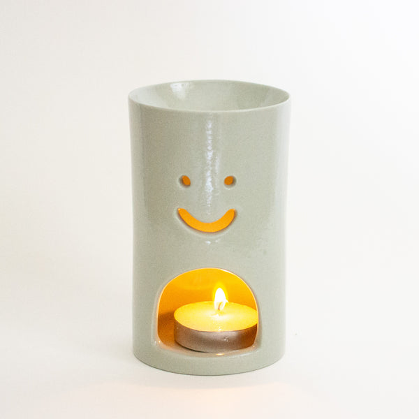 A green ceramic oil burner with a smiling face cutout and a tea light candle burning inside, set against a plain white background.