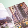 A close-up, angled view of an open book, showing a printed photograph of a city street with tall buildings, overhead wires, and a green streetcar. The foreground page is blurred, and the background around the pages is a pale mint green.