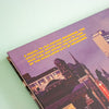 A close-up of a book featuring a vintage-style cityscape in purple tones with bright yellow text, photographed on a pale mint background.