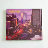 The back cover of a hardcover book, featuring a purple-toned photograph of a city street with long light trails from cars, set against a plain mint background.