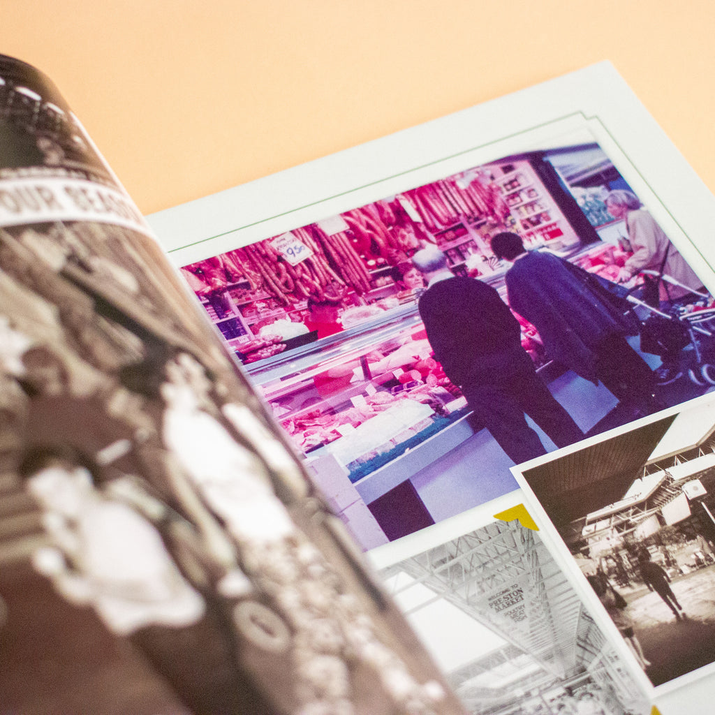 An open printed photo book on a warm beige surface, showing a glossy black-and-white photograph on the left page and a colour photo of a meat market scene on the right page.
