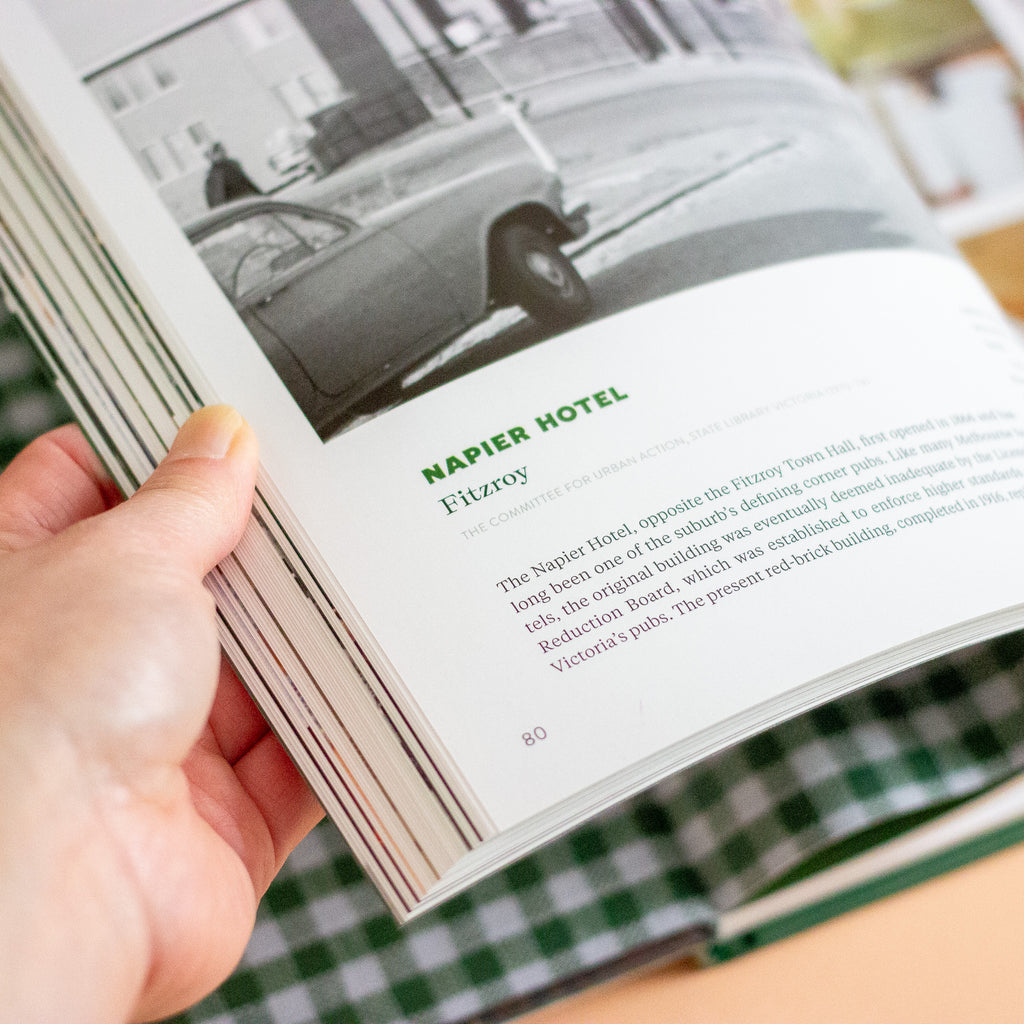 A hand holding an open book or magazine page showing a black-and-white photograph of a car on a street and the text 'NAPIER HOTEL' with a short article below.
