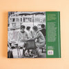 The back cover of a book, shown flat against a solid peach background. The cover features a black and white photograph of women sitting at an outdoor cafe table with a vintage bus in the background, next to a solid green spine band containing white text.