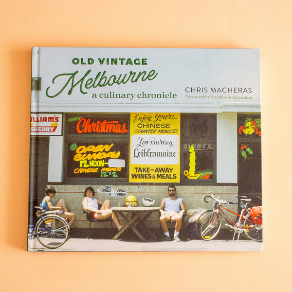 The book 'Old Vintage Melbourne: a culinary chronicle' by Chris Macheras, featuring a colourful cover illustration of a vintage Melbourne street scene with people and bicycles, set against a solid peach background.