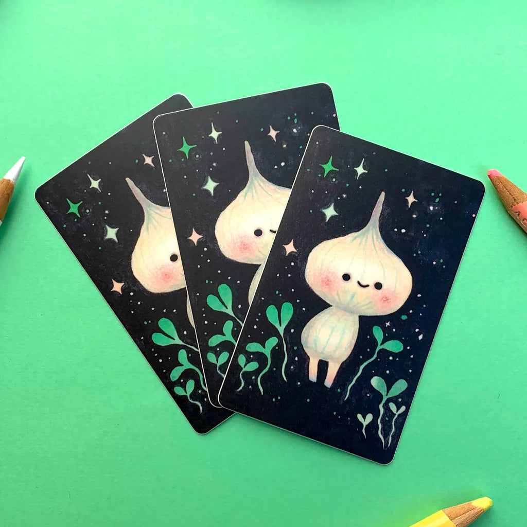 Three illustrated cards with a cute cartoon character on a dark background, arranged on a bright green surface with coloured pencils around them.