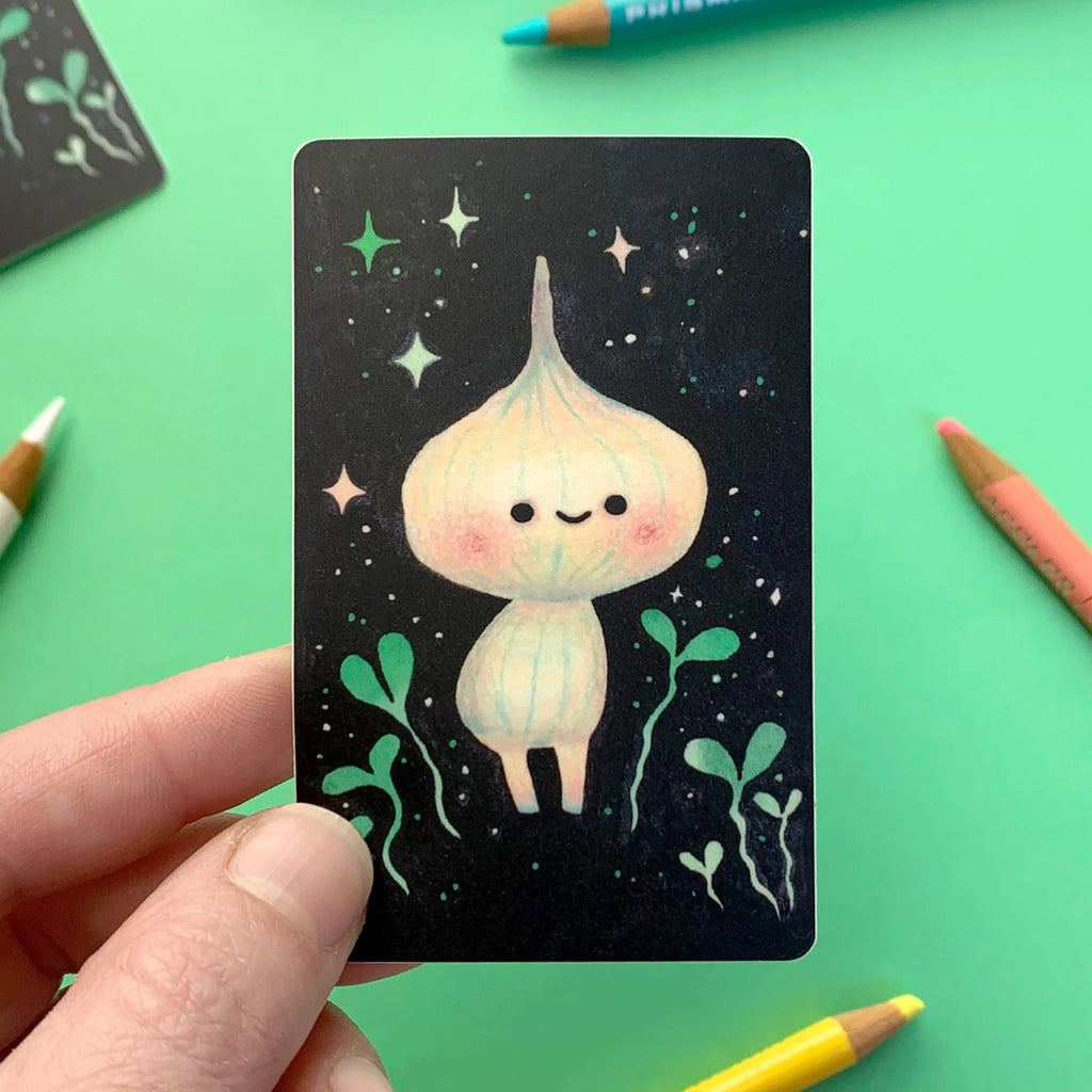 A hand holds a small illustrated card featuring a cute pale character on a black starry background, surrounded by colored pencils on a bright green surface.