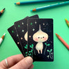 Hand holding a set of cards with a cute character illustration on a green background with pencils.