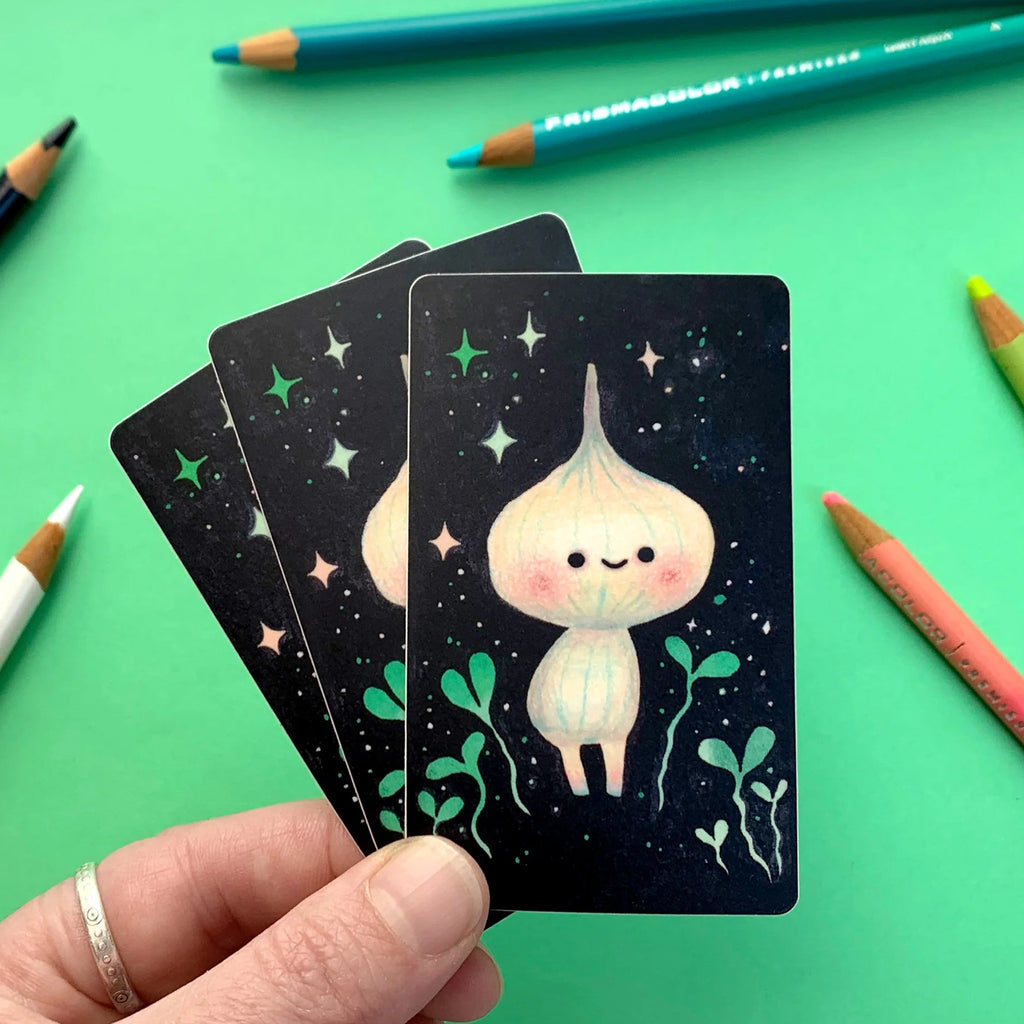 Hand holding a set of cards with a cute character illustration on a green background with pencils.