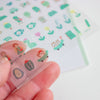 A close-up of a hand holding a transparent sheet of small cartoon stickers sheet. The stickers feature cute illustrated characters, plants, and objects in soft pastel colours including green, pink, yellow, and blue on a white background.