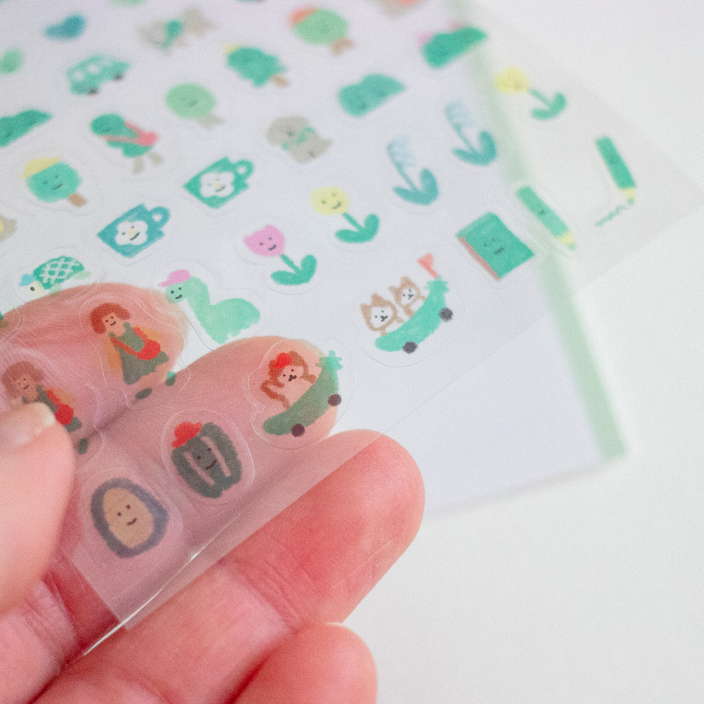 A close-up of a hand holding a transparent sheet of small cartoon stickers sheet. The stickers feature cute illustrated characters, plants, and objects in soft pastel colours including green, pink, yellow, and blue on a white background.