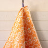 An orange kitchen towel with a white geometric honeycomb pattern and small white flowers with green centers, hanging from a metal hook against a white wood-paneled wall.