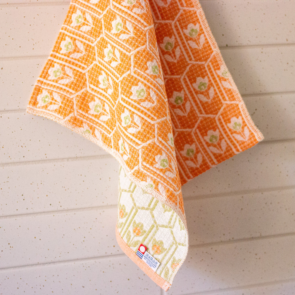 An orange and white patterned kitchen towel with a honeycomb design and small floral motifs, hanging against a white paneled wall.