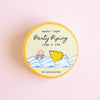 A round roll of washi tape labeled 'Party Piping' by @riomuniandy, featuring a yellow border and a pastel illustration of a yellow cookie and blue waves on a cream background, set against a soft pink backdrop.