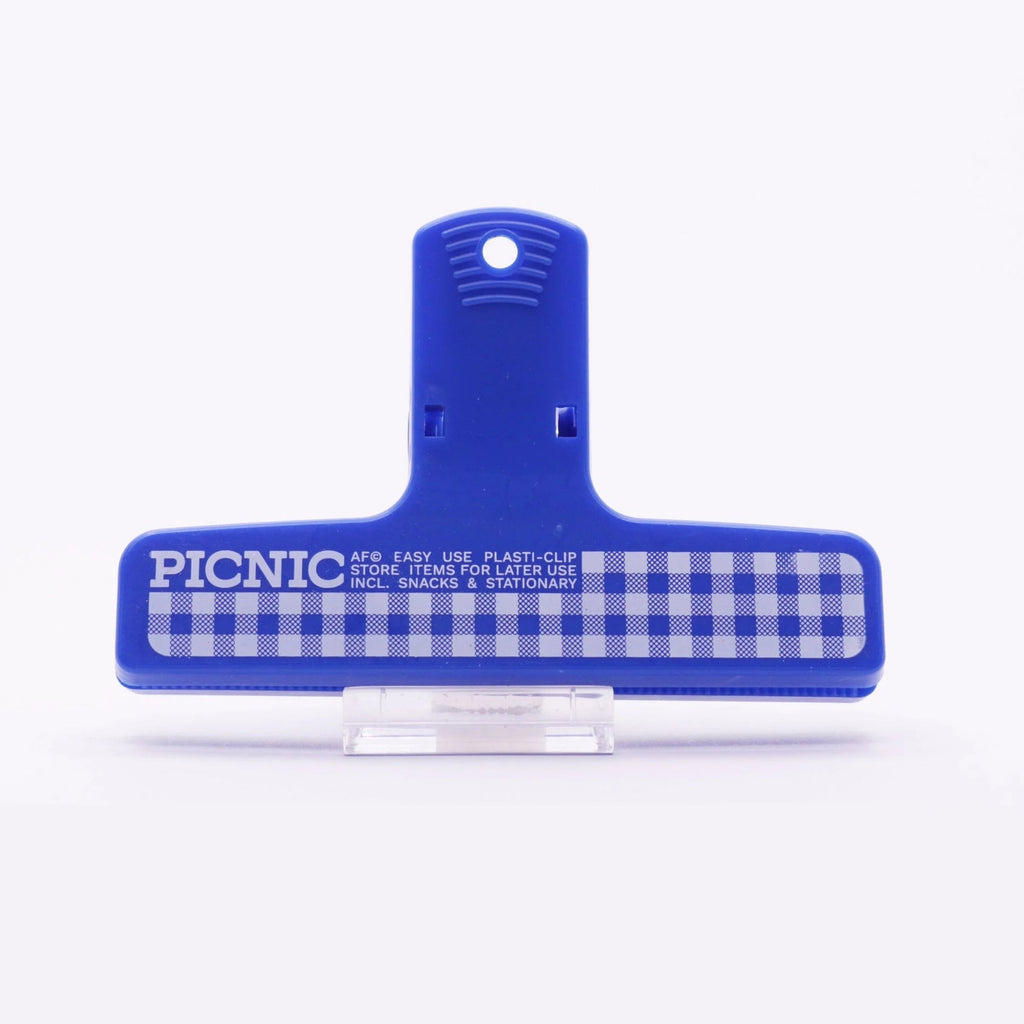 A navy plastic binder clip with white and navy gingham print with text reading 'PICNIC' on the front clipping a cloth, displayed against a white background.