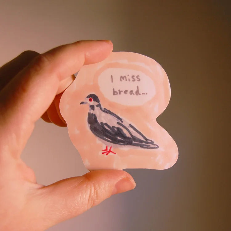 A hand holds a light pink, bread-shaped sticker featuring a black and white bird with a speech bubble that reads 'I miss bread.