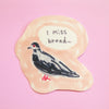 A die-cut sticker featuring a hand-drawn black and white bird with a red eye and red beak, set on a peach-colored background with a speech bubble that reads 'I miss bread.