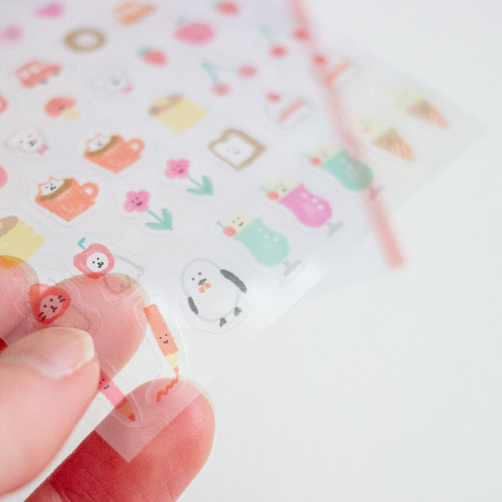 A hand holding a transparent sheet of small pastel stickers with cute illustrations, including a penguin, flowers, and food-themed motifs, against a soft white background.