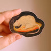 A hand holding an illustrated sticker of a rat eating a slice of pizza with a dark outline. 