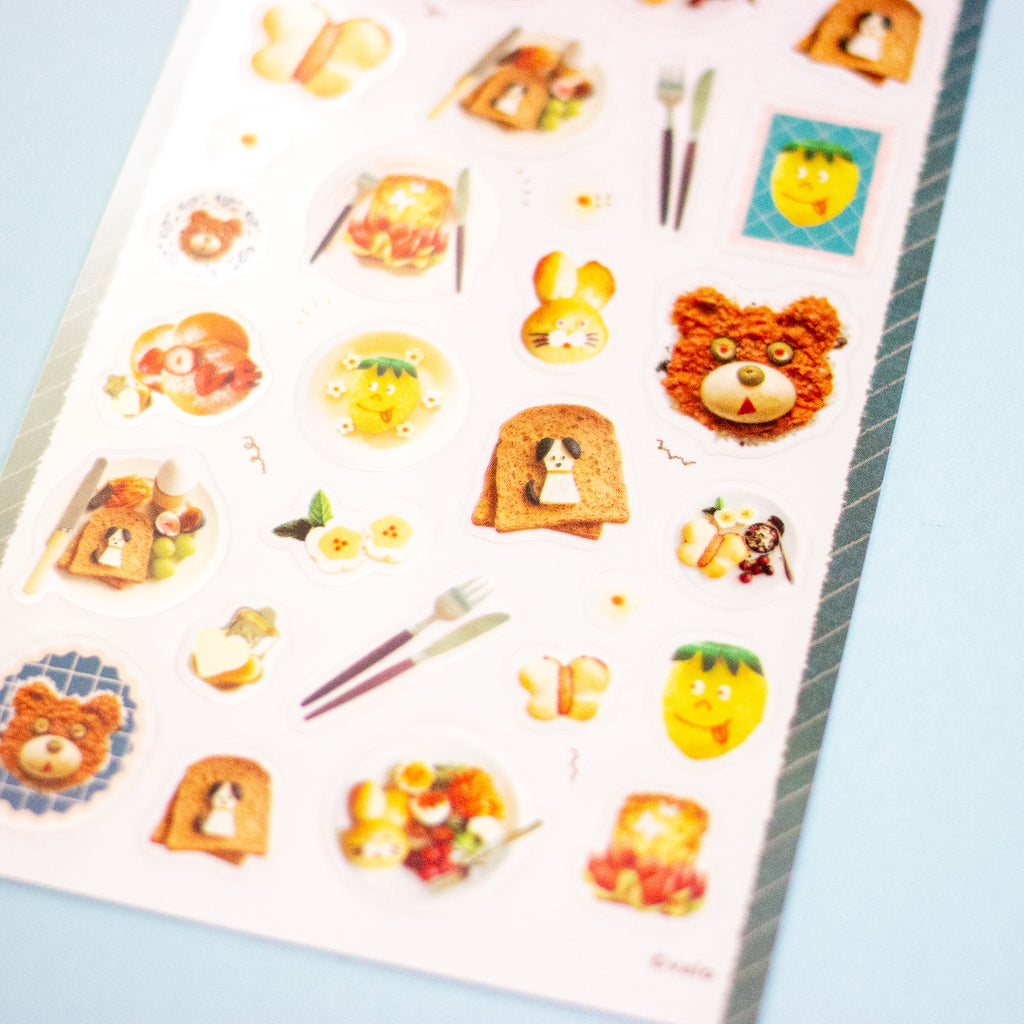 A sheet of cute, food-themed stickers featuring illustrations of toast, breakfast plates, a bear face, and yellow fruit characters, displayed on a light blue background.