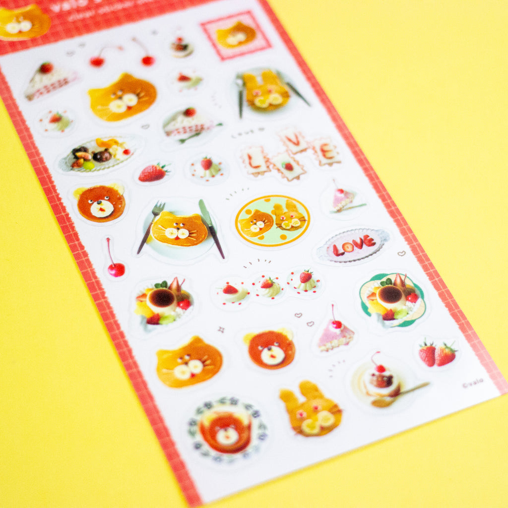 A sheet of cute cartoon stickers featuring yellow cats, strawberries, desserts, cherries, and the word 'LOVE' in letters, placed diagonally on a bright yellow background.