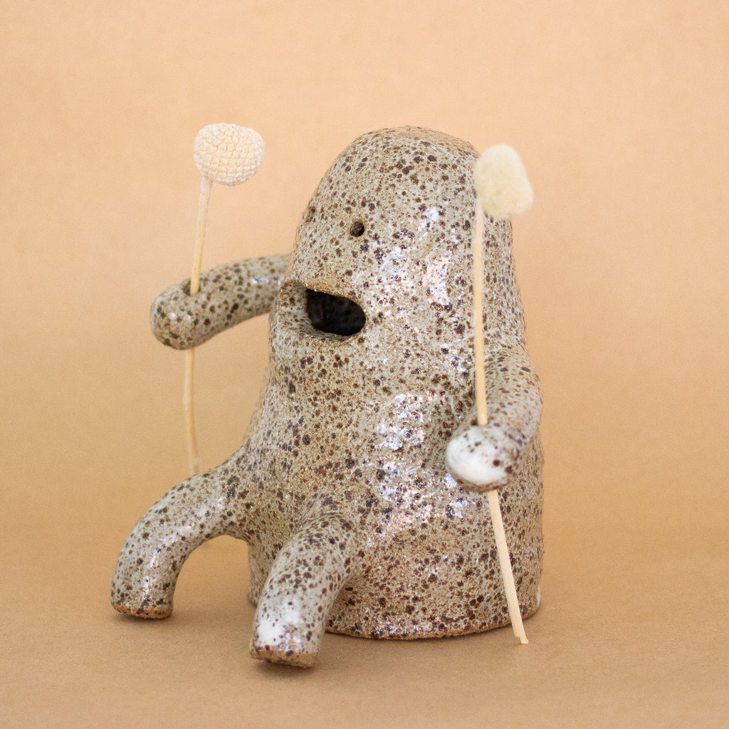 A side view closeup photo of the Plonk handmade stoneware figurine. Sitting down, with two forward facing short legs, and two arms with holes, that are holding two flowers. Figurine has a large mouth opening, and two small eye openings. 