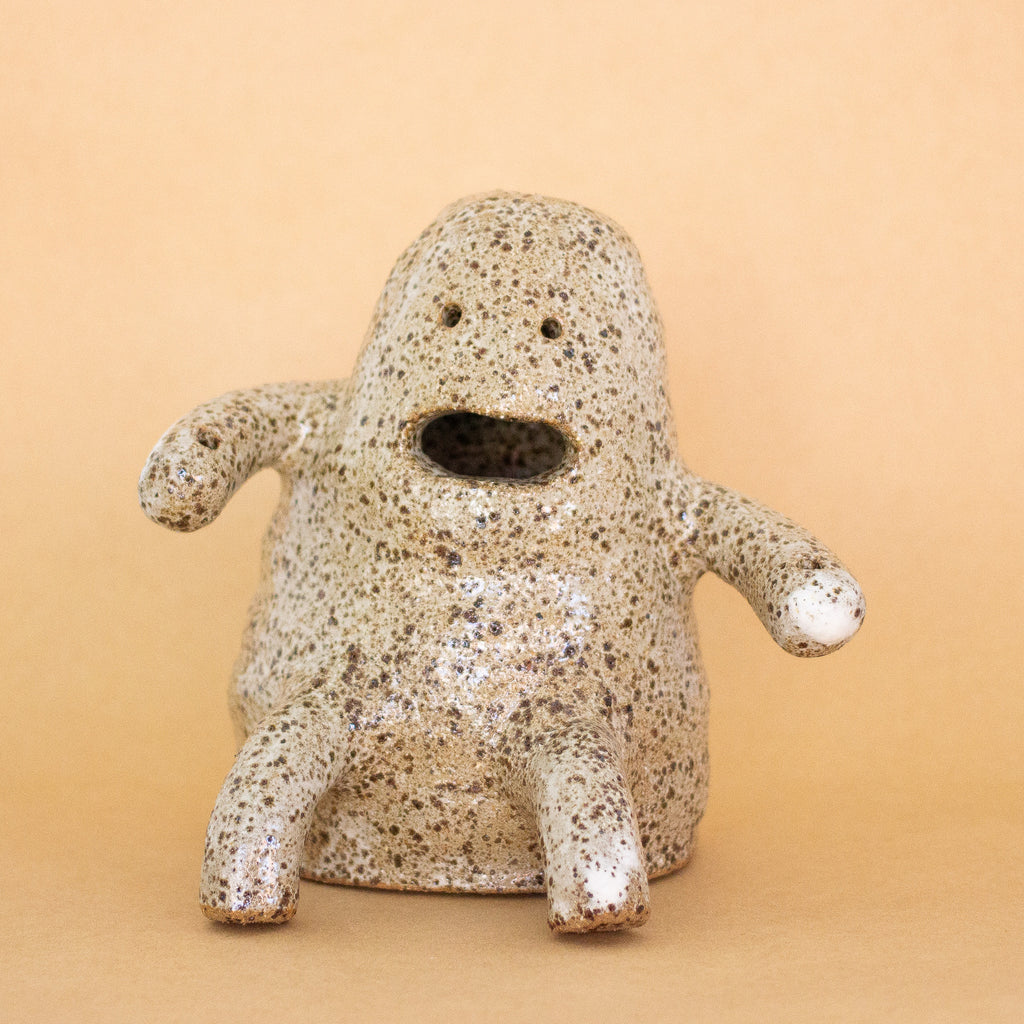 A closeup photo of the Plonk handmade stoneware figurine. Sitting down, with two forward facing short legs, and two arms with holes.  Figurine has a large mouth opening, and two small eye openings. 