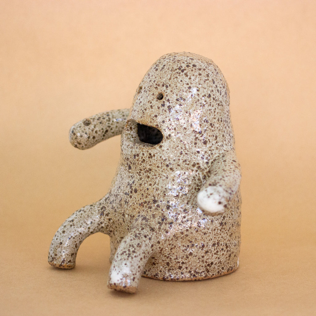 Side view and closeup photo of the Plonk handmade stoneware figurine. Sitting down, with two forward facing short legs, and two arms with holes. Figurine has a large mouth opening, and two small eye openings. 