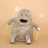 A closeup photo of the Plonk handmade stoneware figurine. Sitting down, with two forward facing short legs, and two arms with holes, that are holding two flowers. Figurine has a large mouth opening, and two small eye openings. 