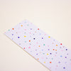 A long rectangular strip of white graph paper with a colourful confetti dot pattern, photographed diagonally on a warm off-white background.