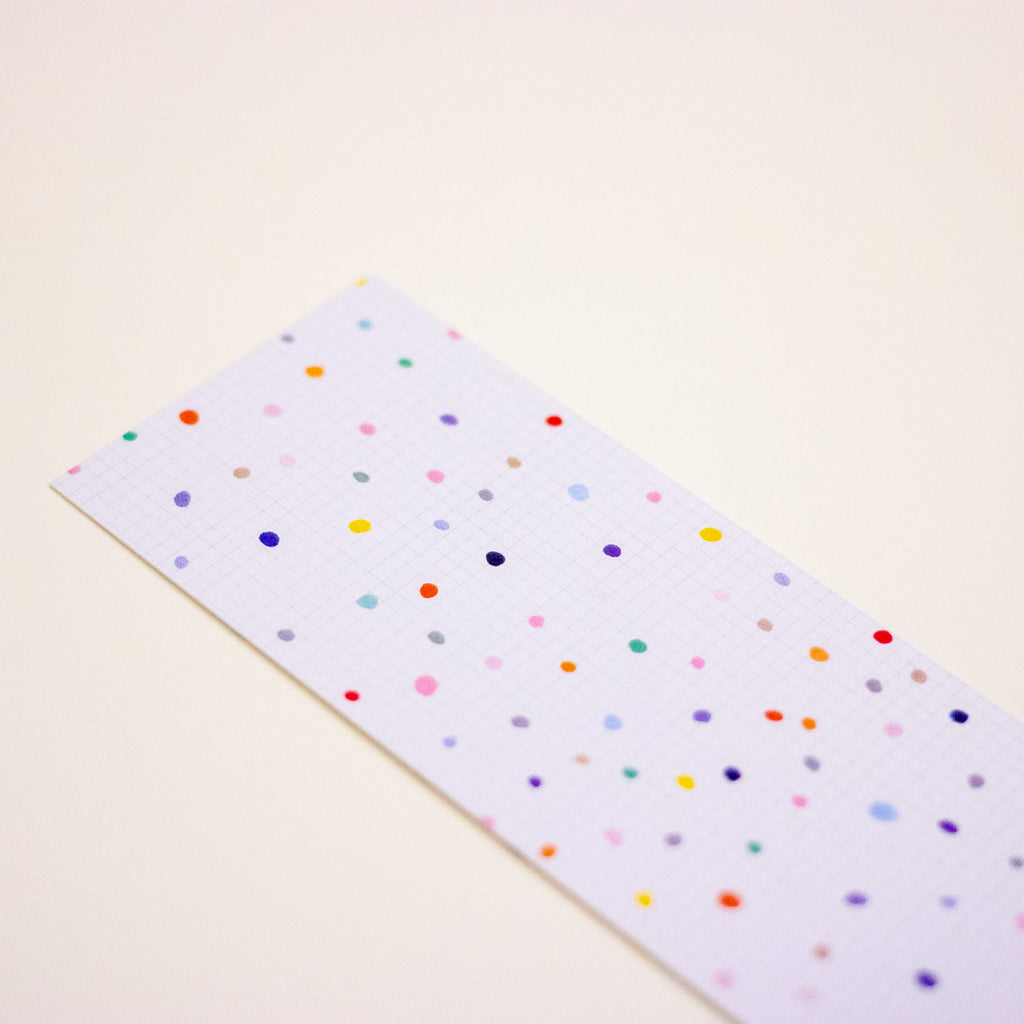 A long rectangular strip of white graph paper with a colourful confetti dot pattern, photographed diagonally on a warm off-white background.