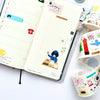 An open planner or journal with decorative stickers and a weekly layout, shown alongside rolls of colourful washi tape on a white surface.