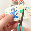 A hand holds a roll of decorative washi tape with a cartoon cat in a blue outfit and small colourful illustrations, while teal-handled scissors cut the tape. Other rolls of tape are blurred in the background.